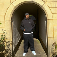 Load image into Gallery viewer, Person wearing a black hoodie and pants standing in an archway.