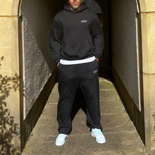 Load image into Gallery viewer, Person wearing a black hoodie and pants with white shoes standing in a stone archway.
